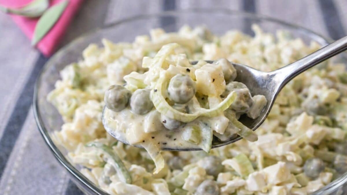 Zbliżenie na srebrną łyżkę z porcją sałatki z porem i groszkiem, prezentujące detale składników: krążki pora, zielony groszek i kostki jajka w gęstym sosie. Sałatka z porem i groszkiem.