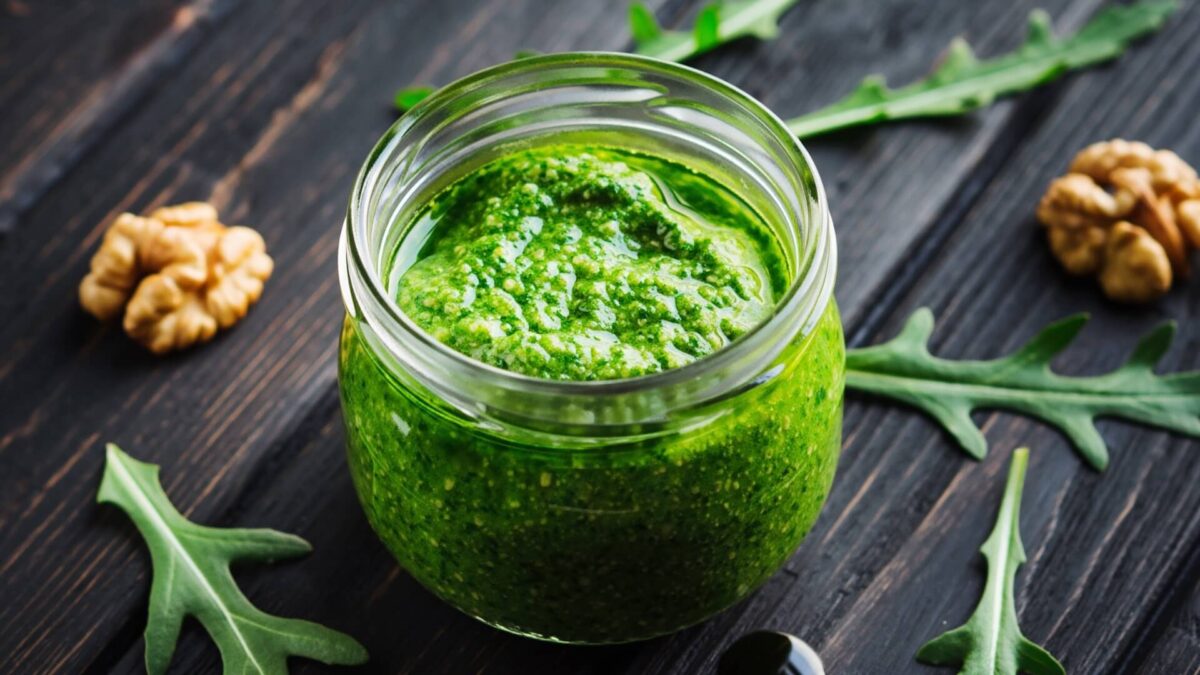 Gotowe pesto z rukoli w szklanym słoiku, udekorowane świeżymi liśćmi i orzechami na ciemnym drewnie.