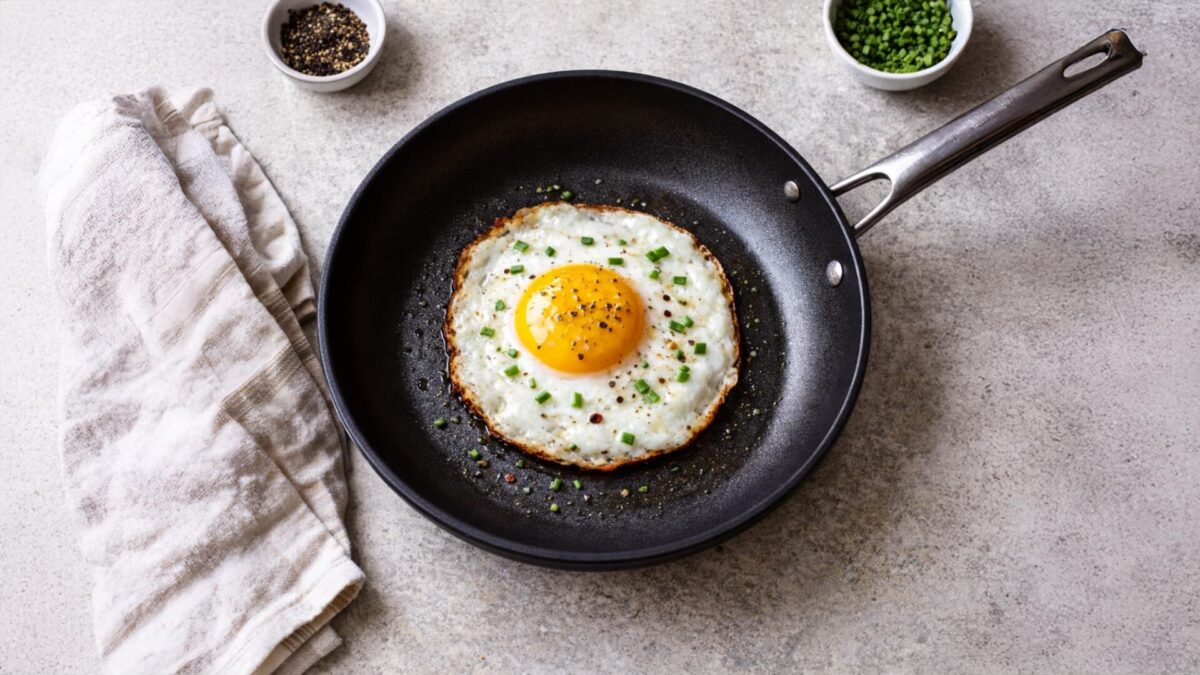 Perfekcyjne jajko sadzone z płynnym żółtkiem, posypane szczypiorkiem i pieprzem, smażone na małej czarnej patelni. Obok lniana ściereczka i miseczki z przyprawami. Widok z góry.