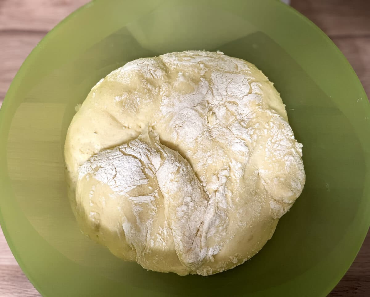 Duża kula jasnożółtego, wyrośniętego ciasta drożdżowego, oprószona mąką, znajdująca się wewnątrz dużej, zielonej plastikowej miski. Widoczne są pęknięcia i struktura ciasta.