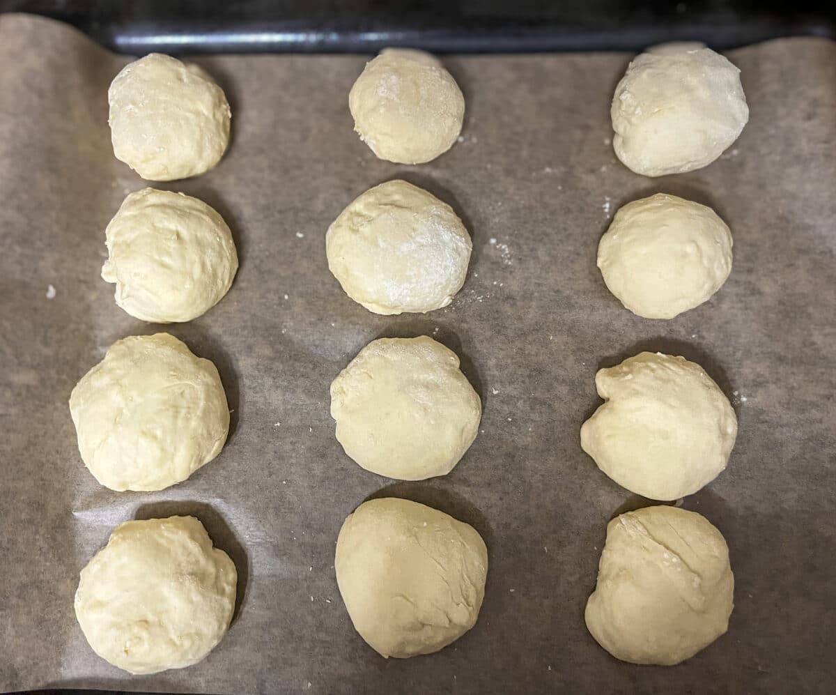 Dwanaście okrągłych kulek surowego ciasta drożdżowego, lekko oprószonych mąką, ułożonych w równych odstępach na blasze do pieczenia wyłożonej brązowym papierem.