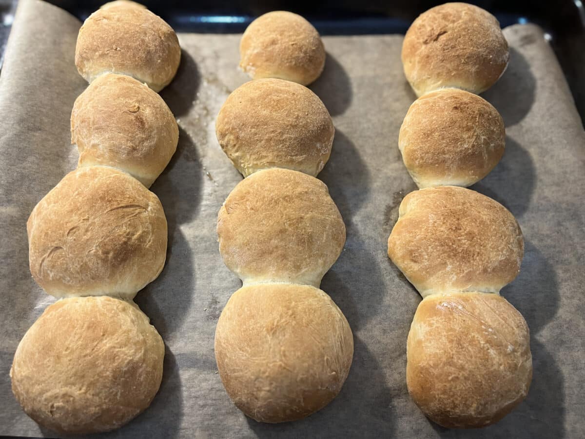 Dwanaście złocistych, okrągłych bułek ułożonych w równych rzędach na blasze wyłożonej papierem do pieczenia. Bułki są rumiane, niektóre stykają się ze sobą, tworząc charakterystyczne połączenia.