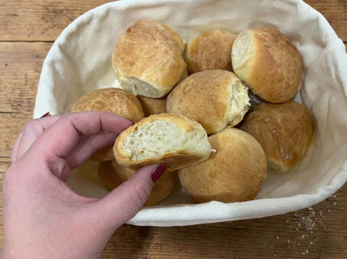 Dłoń trzymająca przekrojoną bułkę, prezentująca jej puszyste, białe i porowate wnętrze na tle drewnianego blatu i koszyka z pieczywem.