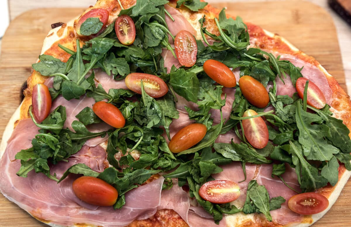 Pizza rzymska z szynką parmeńską i rukolą 1 Zbliżenie na cienką pizzę rzymską z mozzarellą, szynką parmeńską, świeżą rukolą i pomidorkami koktajlowymi.