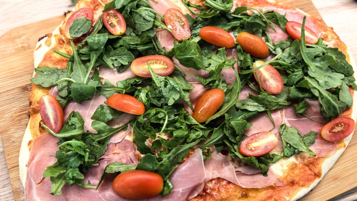 Pizza rzymska z szynką parmeńską i rukolą 3 Pizza rzymska z szynką parmeńską, rukolą i połówkami pomidorków koktajlowych na drewnianej desce.
