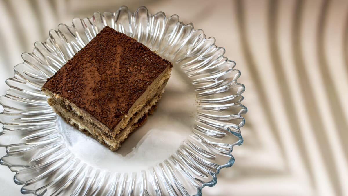 Tiramisu pistacjowe – kremowy włoski deser bez pieczenia 1 Kawałek tiramisu pistacjowego oprószony kakao podany na szklanym talerzu