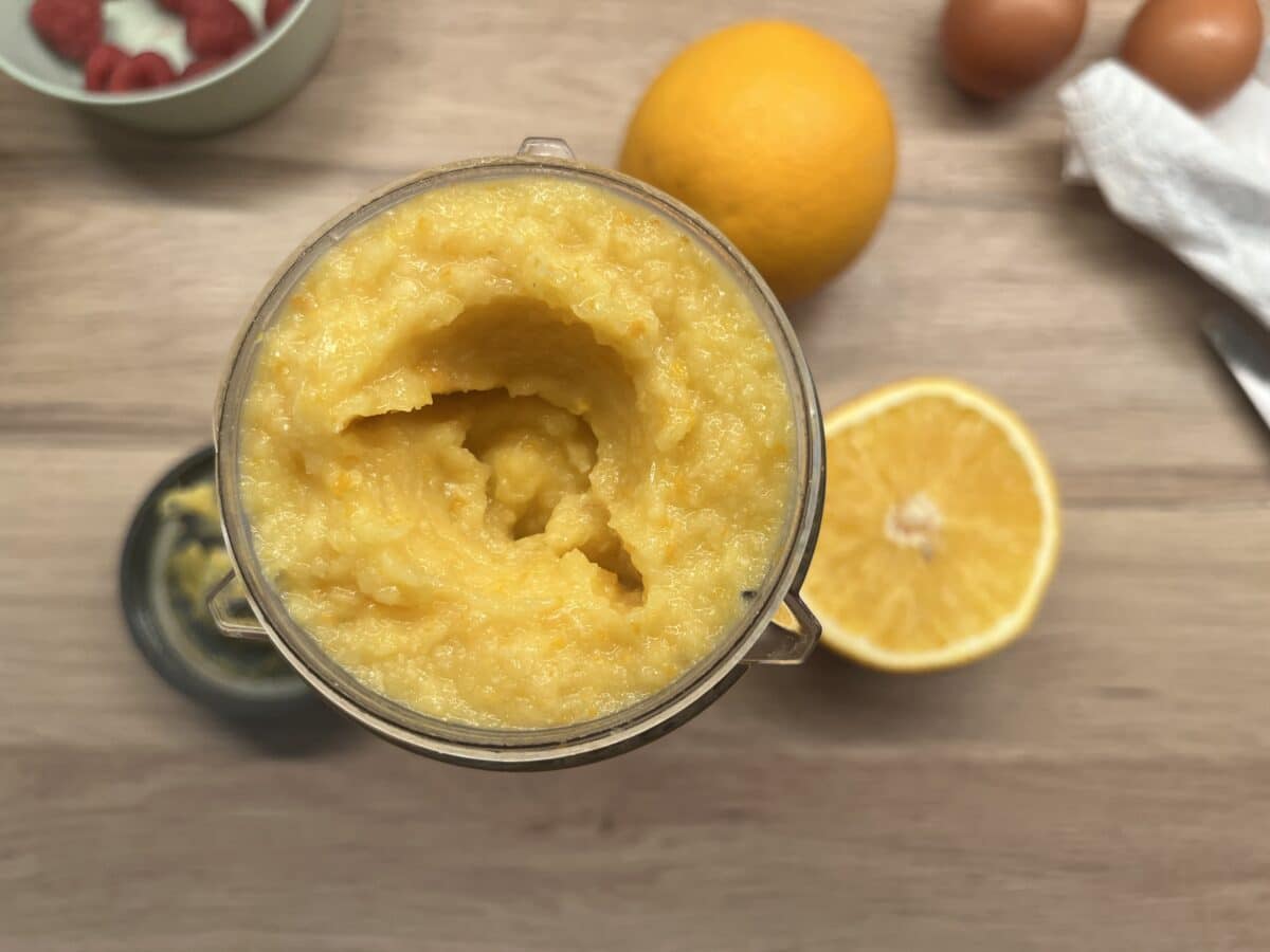 Gładkie puree z gotowanych pomarańczy w pojemniku blendera, obok cała i przekrojona pomarańcza oraz jajka