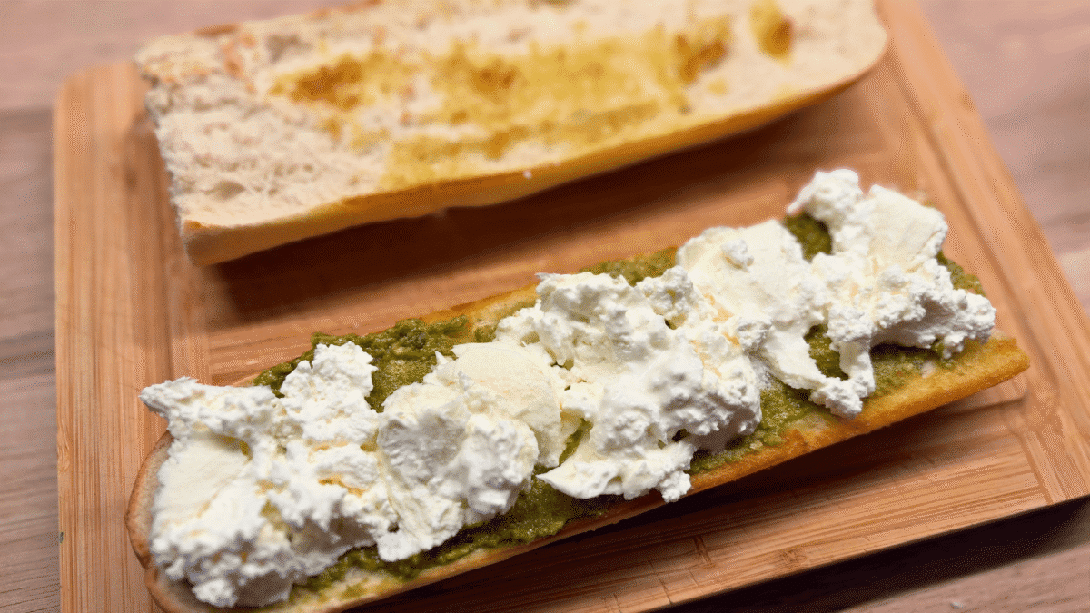 Przekrojona bagietka posmarowana zielonym pesto i przykryta kawałkami burraty.